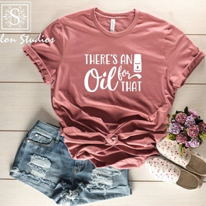 May include: A mauve t-shirt with the text "There's an Oil for That" in white lettering. The shirt is styled with denim shorts and floral patterned shoes.