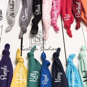 May include: A variety of t-shirt colors displayed on a white wooden surface. The colors include white, athletic heather, storm, black, light pink, charity pink, red, cardinal, coral, lilac, purple, yellow, kelly, royal, navy, turquoise, mint, and sea green.