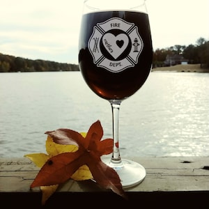 Puede incluir: Una copa de vino con un emblema de bombero en blanco y negro que dice "Fire Dept." y "mother" con un coraz&oacute;n en su interior. La copa est&aacute; llena de vino tinto y est&aacute; sobre una superficie de madera con una hoja marr&oacute;n y amarilla.