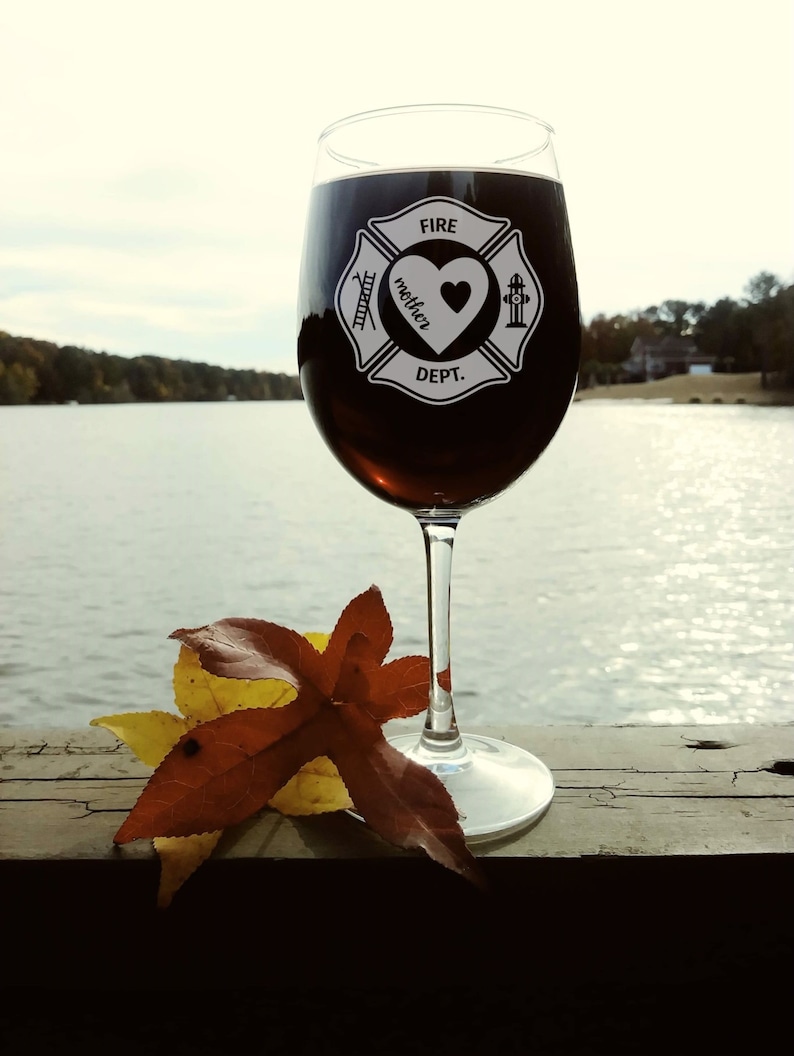 Puede incluir: Una copa de vino con un emblema de bombero en blanco y negro que dice "Fire Dept." y "mother" con un coraz&oacute;n en su interior. La copa est&aacute; llena de vino tinto y est&aacute; sobre una superficie de madera con una hoja marr&oacute;n y amarilla.