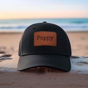 May include: Black baseball cap with a brown leather patch that says "Poppy" on it. The cap is sitting on a sandy surface.