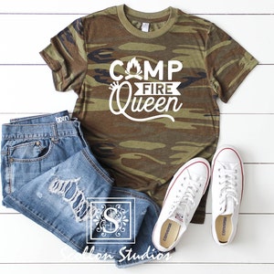 May include: A camouflage green t-shirt with the text "Camp Fire Queen" in white. The shirt is laid flat on a white wooden surface with a pair of white sneakers and blue jeans.