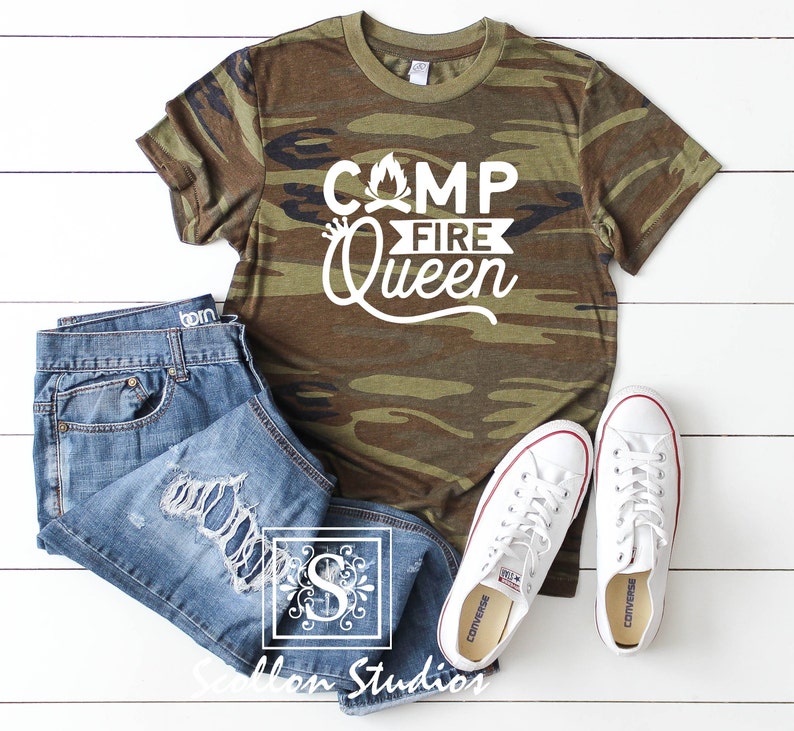 May include: A camouflage green t-shirt with the text "Camp Fire Queen" in white. The shirt is laid flat on a white wooden surface with a pair of white sneakers and blue jeans.
