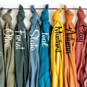 May include: A row of 12 folded t-shirts in various colors, including dust, deep heather, military green, olive, forest, slate, teal, mustard, autumn, clay, maroon, and cardinal. The t-shirts are hanging on a white wooden background.