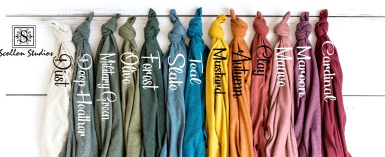 May include: A row of 12 folded t-shirts in various colors, including dust, deep heather, military green, olive, forest, slate, teal, mustard, autumn, clay, maroon, and cardinal. The t-shirts are hanging on a white wooden background.