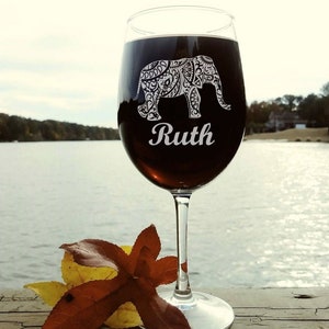 May include: A clear wine glass with a black and white patterned elephant design and the name "Ruth" etched on the side. The glass is filled with red wine and is sitting on a wooden surface with a brown leaf.