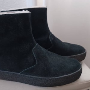 May include: A pair of black suede boots with a short shaft and a rounded toe. The boots have a thick, dark sole and a white, fluffy lining visible at the top. The boots are sitting on a white surface.