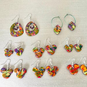 May include: A collection of colorful polymer clay earrings. The earrings feature various shapes, including teardrops, hoops, and Mickey Mouse silhouettes. The earrings are decorated with vibrant patterns of orange, purple, green, and yellow.