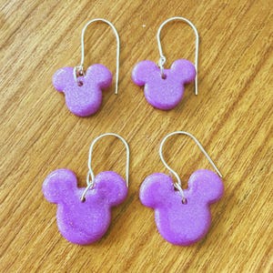 May include: Four pairs of purple Mickey Mouse-shaped earrings with silver-colored hooks. The earrings are a solid, glittery purple color. The earrings are displayed on a wooden surface.
