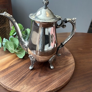 May include: A polished silver-toned metal teapot featuring a tall, slender body, a curved spout with ornate detailing, a matching lid with a decorative finial, and a handle with black accents. The teapot stands on three detailed, ornate feet.