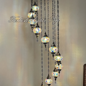 Op de afbeelding: Een meerlaags kroonluchter met 8 kleurrijke glazen bollen die aan een zwart metalen frame hangen. De kroonluchter is 86 inch hoog en 25 inch breed. Elke bol is 5 inch breed en 4 inch hoog. De kroonluchter hangt aan een plafond in een kamer met witte muren.