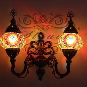 Op de afbeelding: Een wandlamp met twee koppen met sierlijke metalen armen en twee ronde mozaïekglazen bollen. De bollen zijn van binnenuit verlicht en geven een warm licht. De mozaïektegels zijn in tinten blauw, groen, geel en oranje.