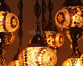 May include: A multi-tiered chandelier with multiple hanging lights. Each light is a round, mosaic-style glass globe with a gold metal frame. The globes are lit and emit a warm, yellow glow. The chandelier is made of black metal and has a decorative design.