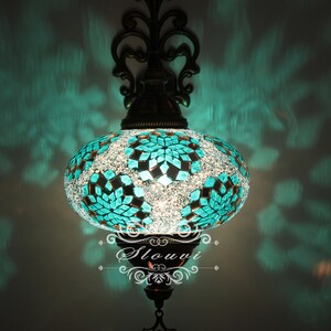 May include: A turquoise mosaic glass wall sconce with a decorative metal frame. The sconce is lit and casting a warm glow.