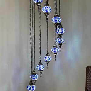 Op de afbeelding: Een spiraalvormige kroonluchter met 9 blauwe en witte mozaïekglazen bollen die aan een zwart metalen frame hangen. De kroonluchter is 86 inch hoog en 25 inch breed. Elke bol is 5 inch breed en 4 inch hoog.