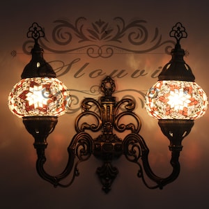 Op de afbeelding: Een wandlamp met twee armen, versierd met sierlijk metaalwerk en twee ronde mozaïekglazen bollen. De bollen zijn gemaakt van kleine, kleurrijke tegels en stralen een warm, uitnodigend licht uit.