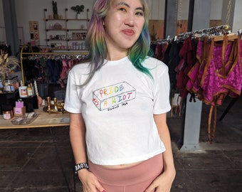 Op de afbeelding: Wit T-shirt met de tekst "Pride Was A Riot" in regenboogkleuren, met "Stonewall 1969" eronder. Het shirt is een cropped model. De persoon draagt een roze legging en een zwarte armband met het woord "love".