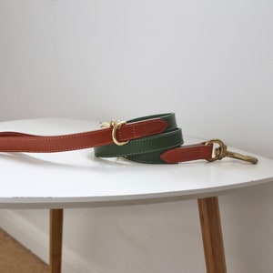 May include: A rolled green and brown leather dog lead with brass fittings. The lead is on a white table. The lead has a handle and a clasp.