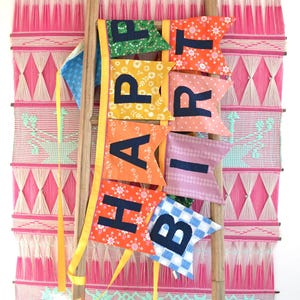 May include: A colorful fabric banner with the words "Happy Birthday" spelled out on individual flags. The banner is hanging on a wooden ladder in front of a pink and white woven wall hanging.