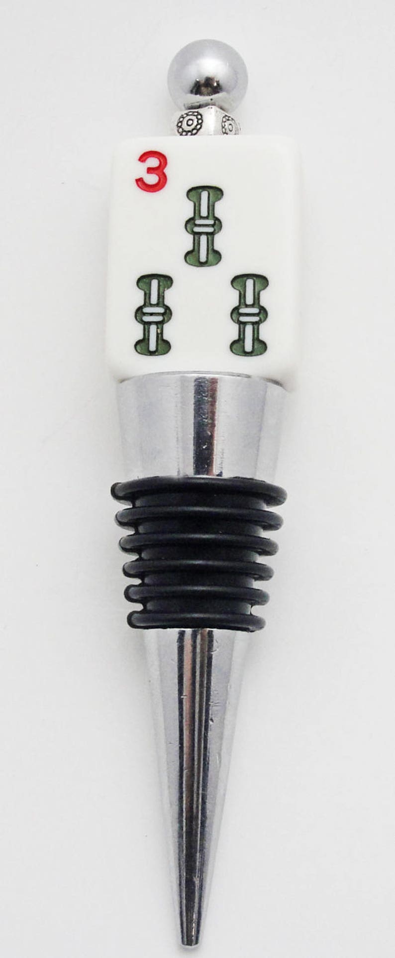 May include: A silver and white bottle stopper with a ceramic top featuring a green and white mahjong tile with the number 3.