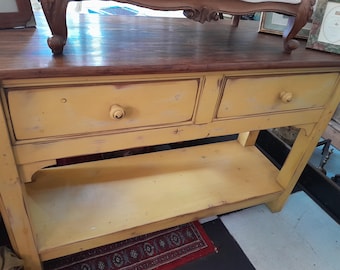 May include: A distressed yellow wooden console table with two drawers and a lower shelf. The top is a dark brown colour. The drawers have round knobs. The table has a rustic, antique appearance.