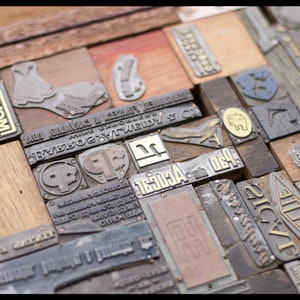 May include: A close-up of a collection of vintage wood and metal printing blocks. The blocks are arranged in a random pattern and feature various designs and text, including "NUMON", "ALL ORNAMENTS & METAL", "RUBBER STAMPS", and "21C/21".