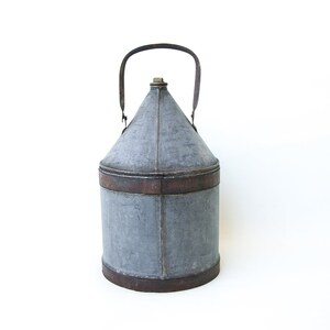 May include: A vintage galvanized metal jug with a leather handle. The jug has a conical top and a cylindrical base. The jug is weathered and shows signs of age.