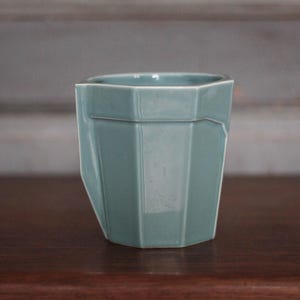 May include: A teal-colored, faceted ceramic cup or small planter. The vessel has a geometric design with angled sides and a slightly irregular top edge. It sits on a dark wooden surface.