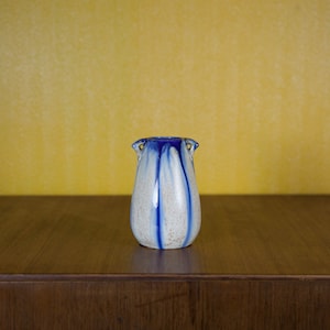 Peut inclure: Un petit vase en céramique blanche avec un glaçage bleu et deux anses. Le vase a une texture mouchetée et un corps en forme de poire.