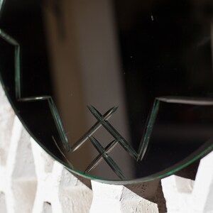 May include: A round mirror with a decorative, geometric pattern etched into the glass. The mirror is framed in a thin, black metal frame.