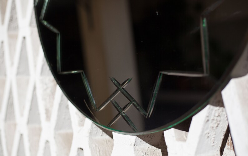 May include: A round mirror with a decorative, geometric pattern etched into the glass. The mirror is framed in a thin, black metal frame.