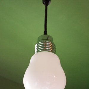 May include: A white light bulb with a silver metal base hangs from a black cord against a green wall.