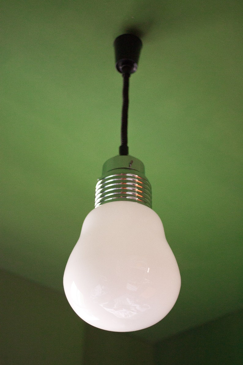 May include: A white light bulb with a silver metal base hangs from a black cord against a green wall.