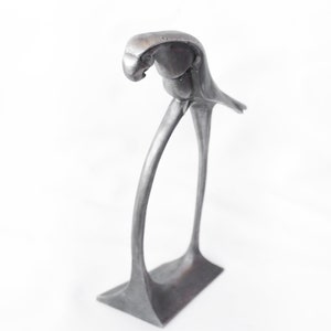 Op de afbeelding: Een zilveren metalen papegaaibeeldje dat op een gebogen voet staat. De papegaai zit op &eacute;&eacute;n poot met zijn kop naar beneden gekanteld.