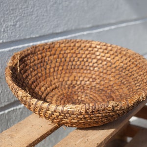 May include: A large, round, woven basket made of natural materials. The basket is brown and has a natural, rustic look. It is sitting on a wooden surface.