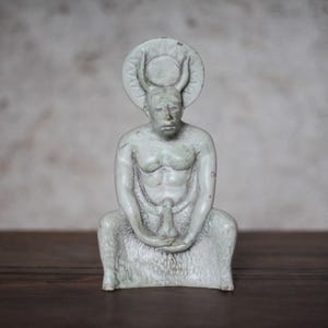 May include: A pale, off-white ceramic figurine depicting a seated figure with horns and a circular halo-like disc behind the head. The figure has a muscular torso, textured lower body, and is holding a phallic object in the lap area.