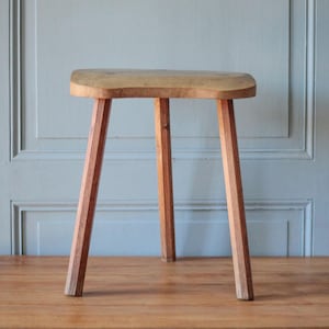 Op de afbeelding: Een houten kruk met drie poten, voorzien van een lichtbruine, gladde, ronde zitting en drie rechte, taps toelopende poten. De kruk is gemaakt van natuurlijk hout met zichtbare nerven en heeft een eenvoudig, functioneel ontwerp met een stevige basis.