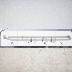 May include: A white ceramic wall-mounted towel rack with a chrome trim and a silver metal bar. The rack has six hooks, four on the bottom and two on the top.