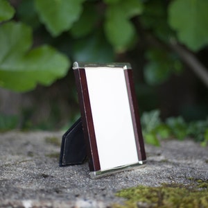 May include: A small, rectangular picture frame with a white interior and a dark brown wooden border. The frame has a silver-colored trim and a black stand. The background is blurred, with green foliage and a stone surface.