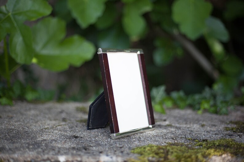 May include: A small, rectangular picture frame with a white interior and a dark brown wooden border. The frame has a silver-colored trim and a black stand. The background is blurred, with green foliage and a stone surface.
