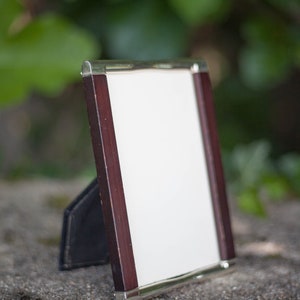 May include: A rectangular picture frame with a white interior, dark wood sides, and silver metal trim. The frame is propped up on a black stand, set against a blurred green background.