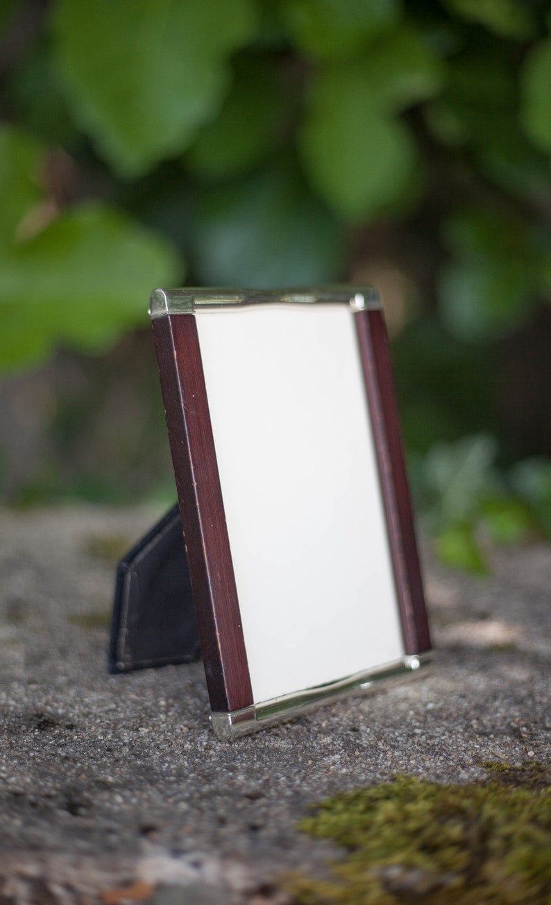 May include: A rectangular picture frame with a white interior, dark wood sides, and silver metal trim. The frame is propped up on a black stand, set against a blurred green background.