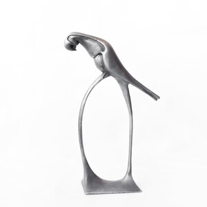 Op de afbeelding: Een zilveren metalen sculptuur van een vogel die op een gebogen standaard zit. De vogel is in een gestileerde art deco-stijl.