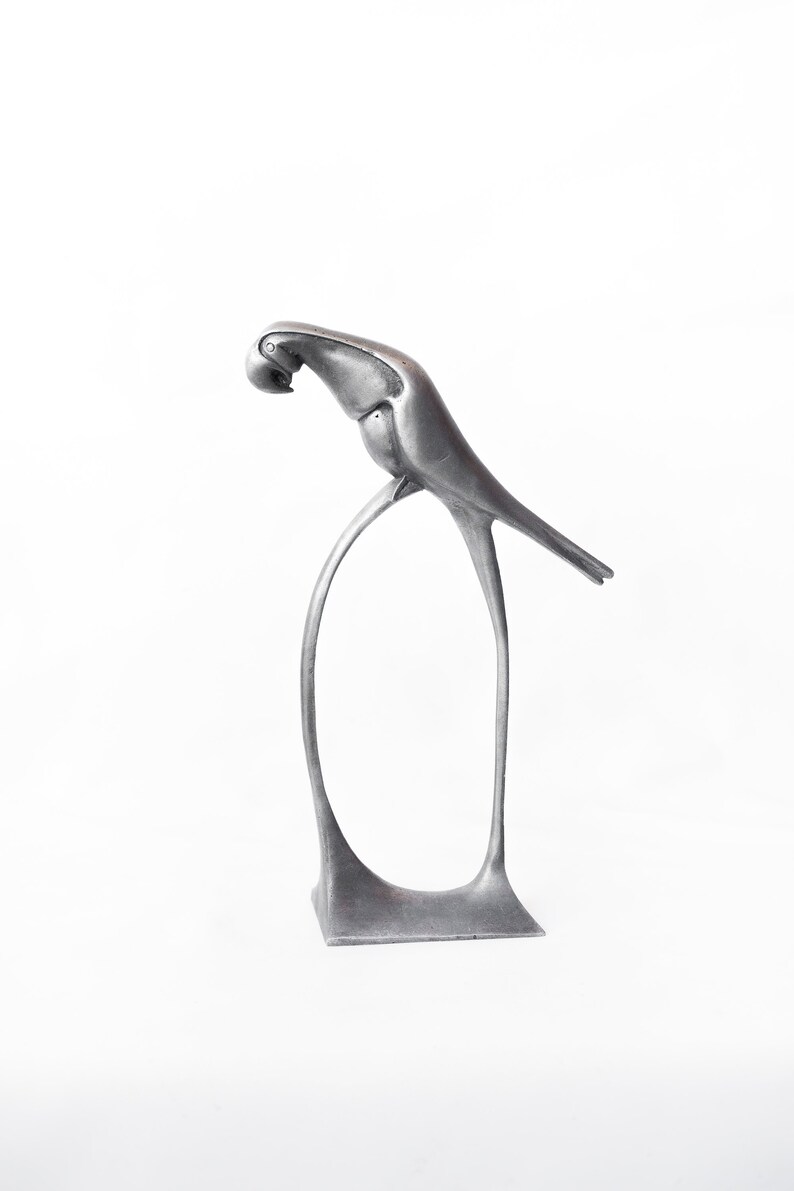 Op de afbeelding: Een zilveren metalen sculptuur van een vogel die op een gebogen standaard zit. De vogel is in een gestileerde art deco-stijl.
