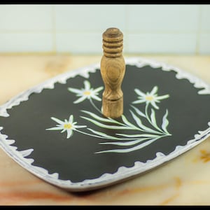 Könnte beinhalten: Ein schwarzes und weißes Keramik-Serviertablett mit einem weißen gewellten Rand. Das Tablett zeigt ein stilisiertes Blumenmuster mit weißen Blüten und grünen Blättern. Ein Holzgriff befindet sich in der Mitte des Tabletts.