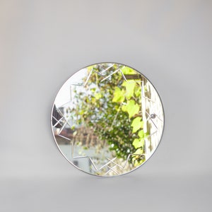 May include: A round mirror with a geometric design etched into the glass. The mirror reflects a view of green foliage and a building in the distance.