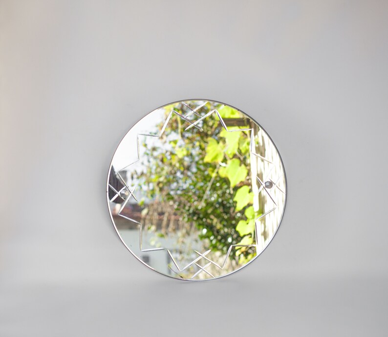May include: A round mirror with a geometric design etched into the glass. The mirror reflects a view of green foliage and a building in the distance.