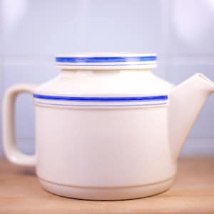 May include: Cream-colored ceramic teapot with a lid and a handle. The teapot is adorned with two thin, parallel blue stripes around the body and lid. The teapot is sitting on a wooden surface.