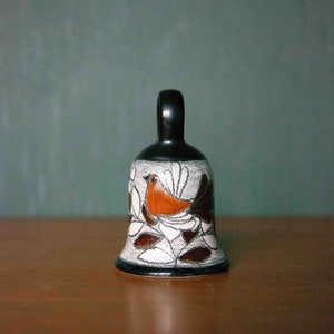 May include: A ceramic bell with a black handle and a white and brown design featuring a bird with orange and brown feathers. The bell is decorated with white flowers and leaves.
