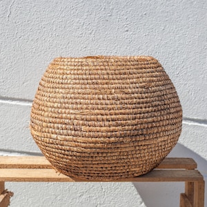 May include: A large, round, woven basket made of natural materials. The basket is brown and has a smooth, textured surface. It is sitting on a wooden shelf.